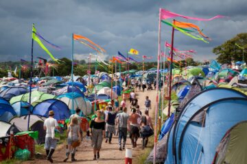 Camping festival rempli de tentes colorées avec des festivaliers marchant sur un chemin, sous un ciel nuageux et décoré de fanions colorés.